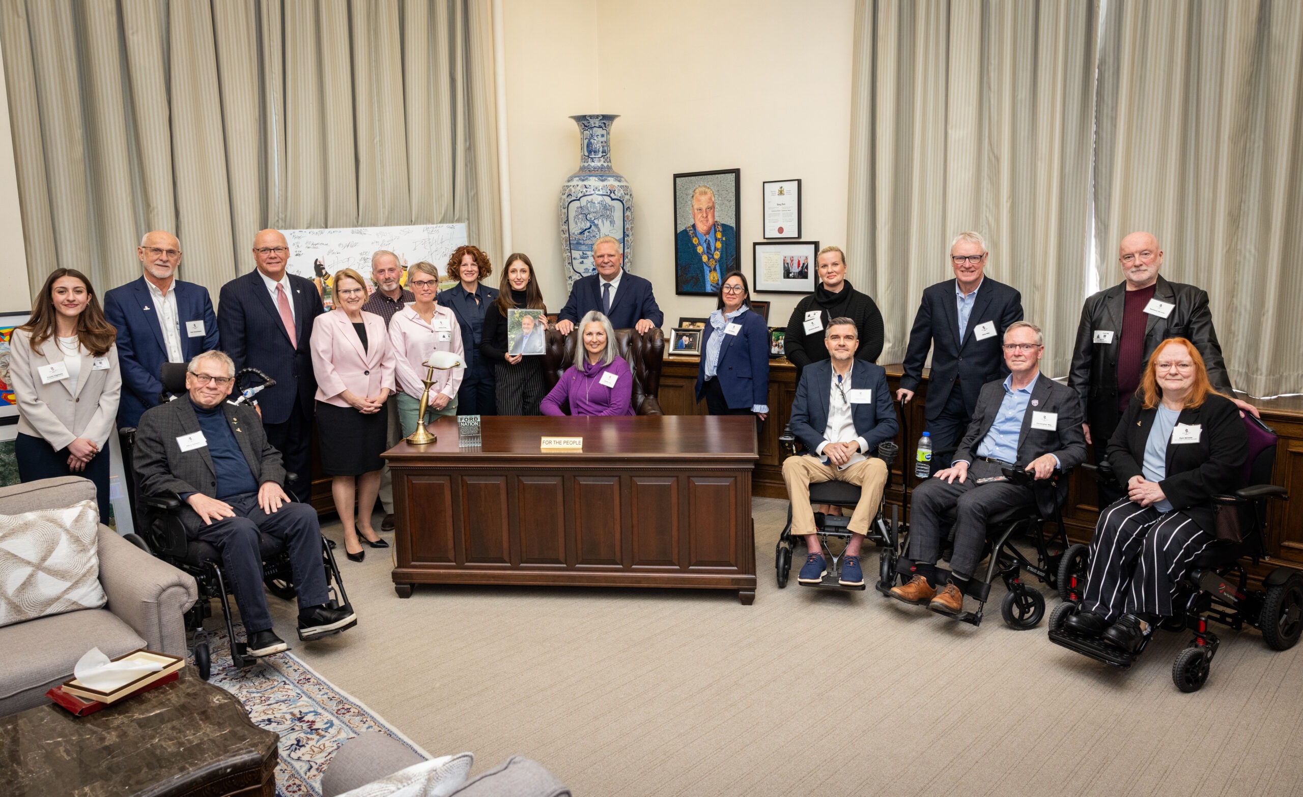 Group of officials and staff posing for a formal group photo in a government chamber around a large wooden desk.
