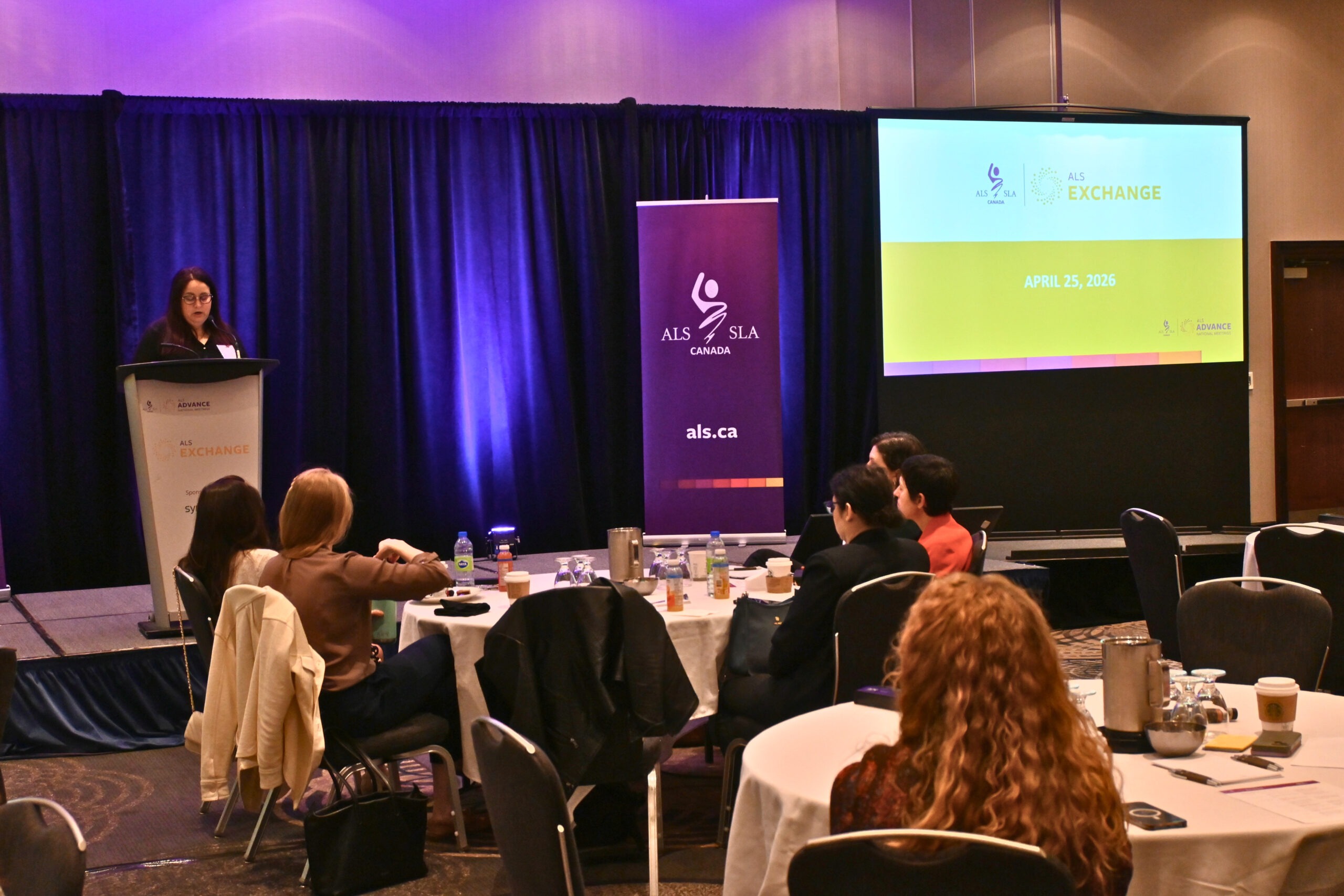 Female speaker stands at a podium on a stage, presenting to an audience in a conference room with a large screen showing ALS Exchange branding and a date.